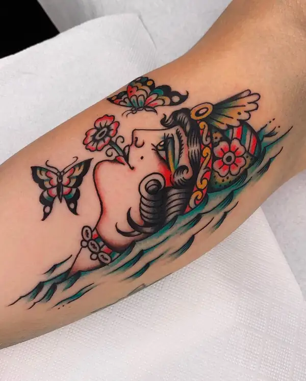 Cabeza de mujer sobre las olas, con flores en el pelo y dos mariposas, en tradicional a color en el brazo.