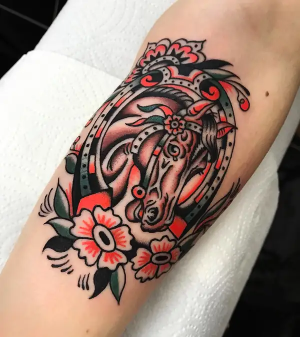 Cabeza de caballo dentro de una herradura, con flores rojas, en tradicional a color en el brazo.