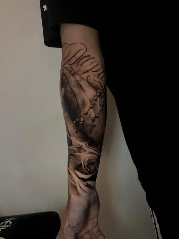 Manos rezando con un rosario y una rosa, en negro y grises en el antebrazo.