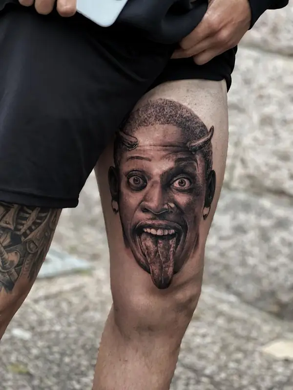 Retrato de un hombre con cuernos de diablo que saca la lengua, en negro y grises en el muslo.