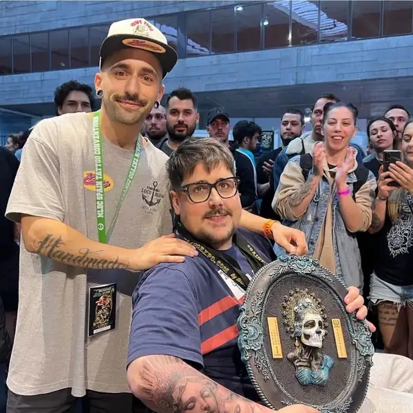 Haroz con su cliente, sentado y con la placa del primer premio, entre el público de una convención de tatuaje.