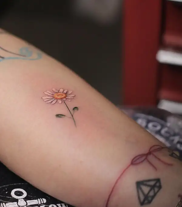 Una margarita a color, un hilo rojo y un diamante en línea fina, en el brazo.