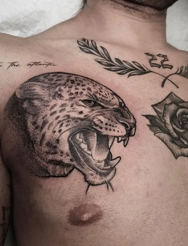 Cabeza de leopardo rugiendo en puntillismo en el pecho, junto a una rosa y una rama de olivo.
