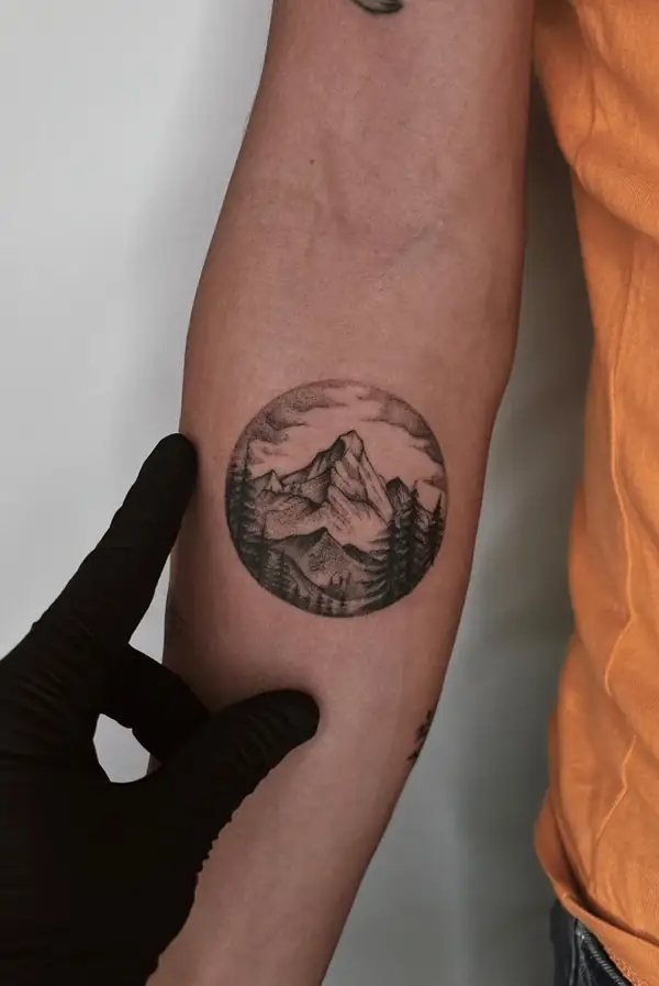 Montañas y pinos dentro de un círculo, en línea fina y punteado en el antebrazo.