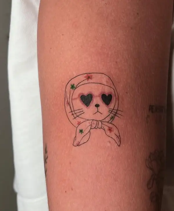 Un gato con pañuelo y ojos de corazón, en línea fina con toques de color, en el brazo.