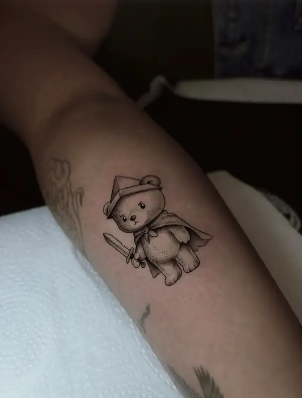 Un osito de peluche con capa y espada, en puntillismo en el antebrazo.