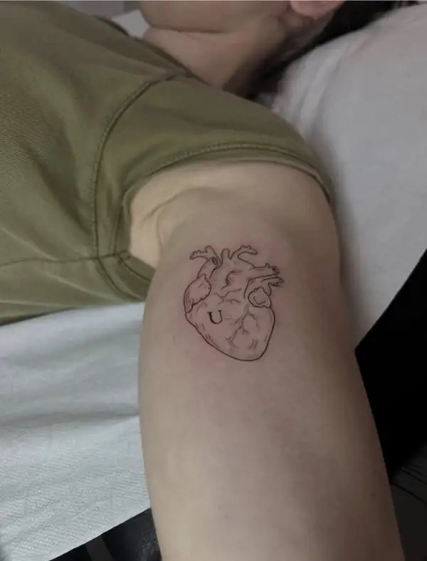 Un corazón anatómico en línea fina con una letra dentro, en el brazo.