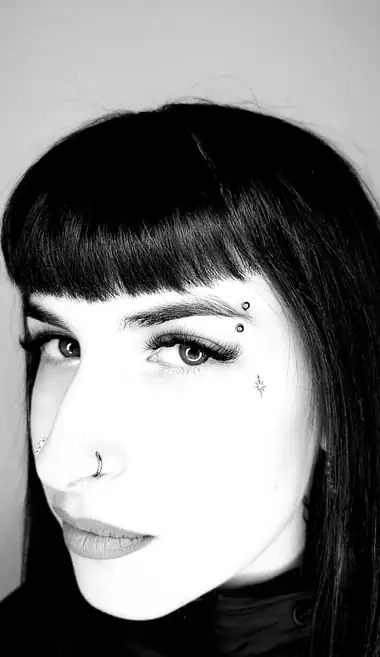 Chica con flequillo, con una barra en la ceja, un aro en la nariz y una estrella pequeña bajo el ojo, en blanco y negro.